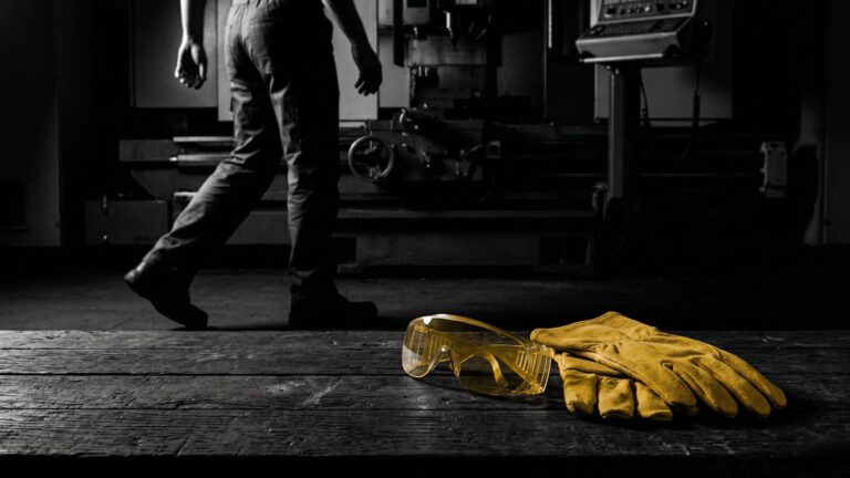 Gefahren durch fehlende Schutzausrüstung im Betrieb: Eine ausdrucksstarke Schwarz-Weiß-Aufnahme von ungenutzter PSA auf einer Werkbank, hervorgehoben durch den gold-gelben Impulspfeil der Unternehmensberatung Christian Harrer, um das Bewusstsein für die Safety Culture zu schärfen.