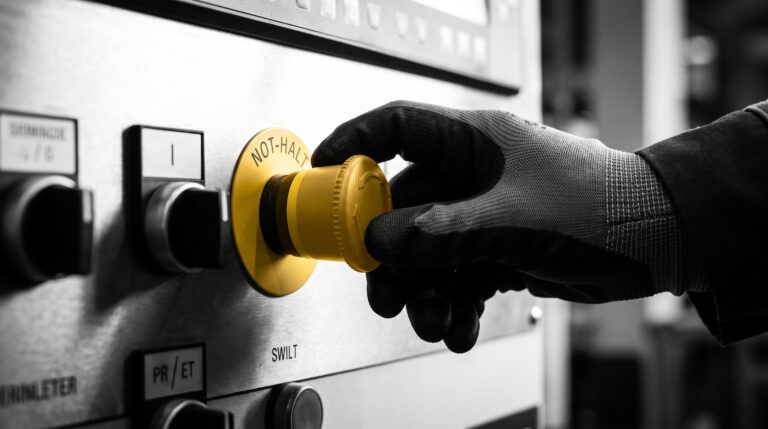 Anerkennung für Arbeitsschutz: Eine kontrastreiche Schwarz-Weiß-Aufnahme einer Hand, die einen in CH-Goldgelb hervorgehobenen Not-Halt-Taster drückt, als Symbol für mutiges und sicheres Eingreifen im Betriebsalltag.